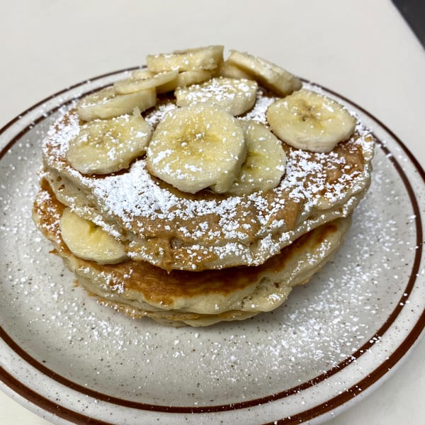 Stack of pancakes topped with sliced bananas, peanut butter, and powdered sugar on a speckled plate.