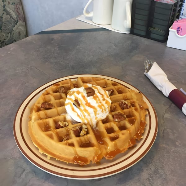 A golden waffle topped with whipped cream, caramel drizzle, and walnuts on a plate at Zorbas Family Dining.