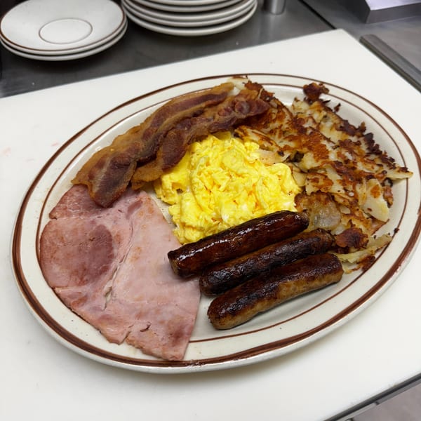 A large breakfast platter with scrambled eggs, bacon, ham, sausage links, and hash browns.